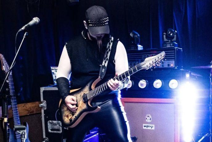 Rock guitarist performing onstage in dark outfit with glowing blue lights and amps behind him.
