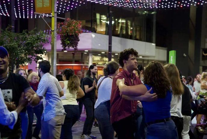 Crowd dancing outdoors under colorful lights with pairs moving energetically in a lively street scene.