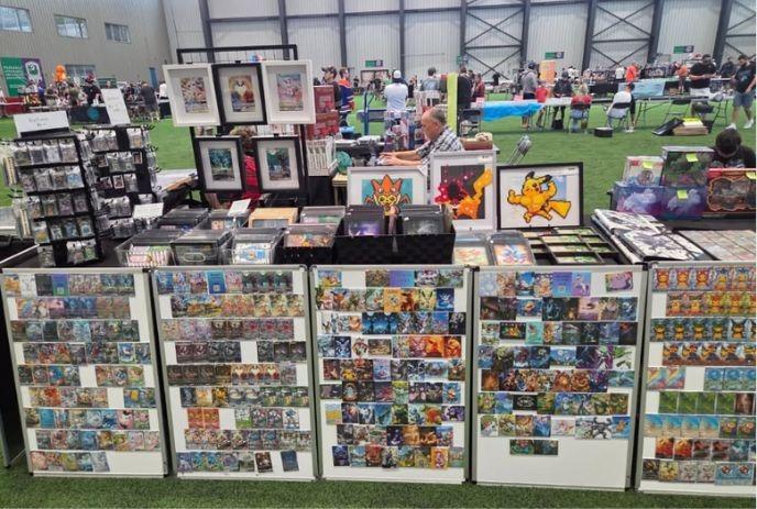 Vendor booth with colorful trading cards, framed art, and people browsing at indoor convention.