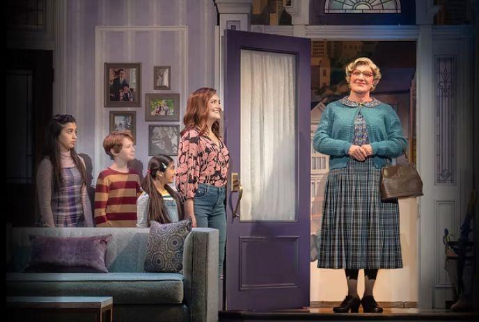 Stage scene with group by couch greeting person entering in plaid dress teal cardigan glasses and holding handbag.
