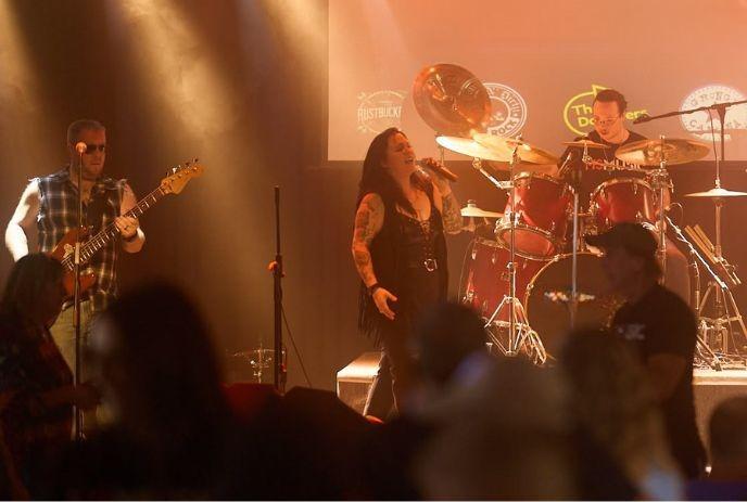 Singer with mic fronts guitarist and drummer on warmly lit stage as audience watches energetic performance.