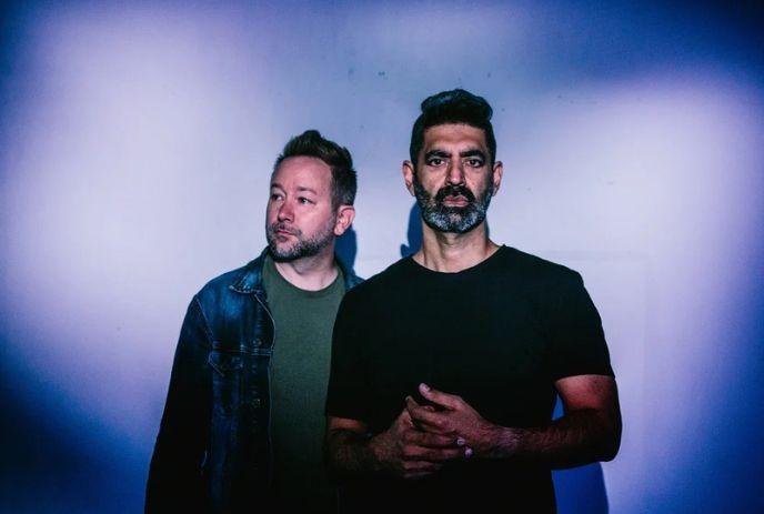 Two artists standing against blue purple backdrop wearing denim jacket and black shirt in moody lighting.