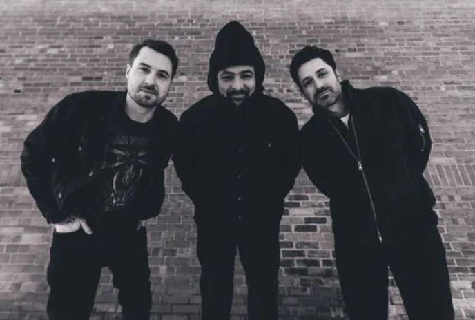 Three artists leaning forward in black and white portrait standing before brick wall in close formation.