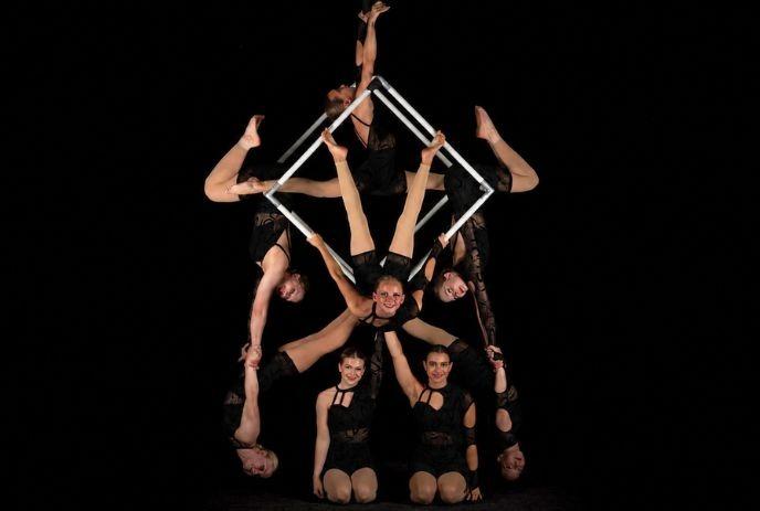 Six acrobats forming balanced geometric pose with inverted performers holding square frame on dark stage.