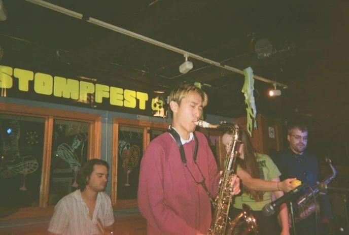 Four musicians perform indoors under Stompfest sign with saxophones keyboard and warm cozy bar lighting.