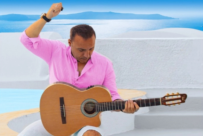 Artist in bright pink?shirted person sitting outdoors by white Mediterranean architecture, holding a guitar with blue sea .