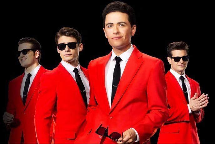 Four artists in red suits white shirts and black ties stand against dark background three wear sunglasses one holds them.
