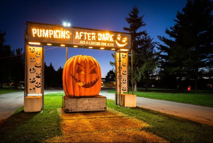 A large carved pumpkin sits under a Pumpkins After Dark sign on a dark evening.