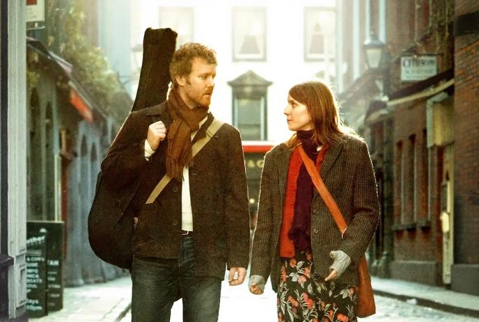 Two people walking down narrow cobblestone street with warm sunlight buildings lining both sides calmly