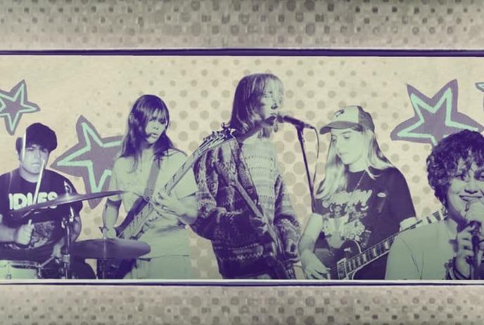 Five musicians perform together with guitars bass drums and singer before bold star patterned backdrop.