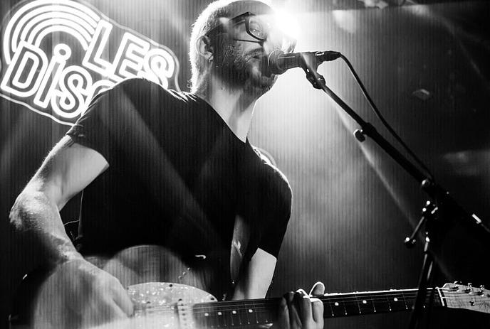 A black and white photo of Eric Howden singing on stage with a guitar.