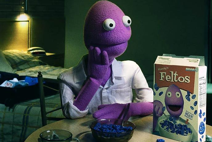 Purple puppet with wide eyes sits pensively at a table with blue cereal and a matching box.