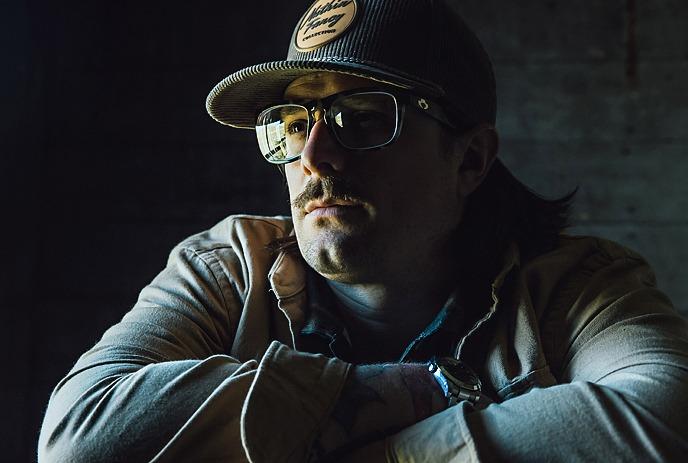Hardy wearing a cap and glasses leans on a chair, illuminated by dramatic side lighting on a dark background.