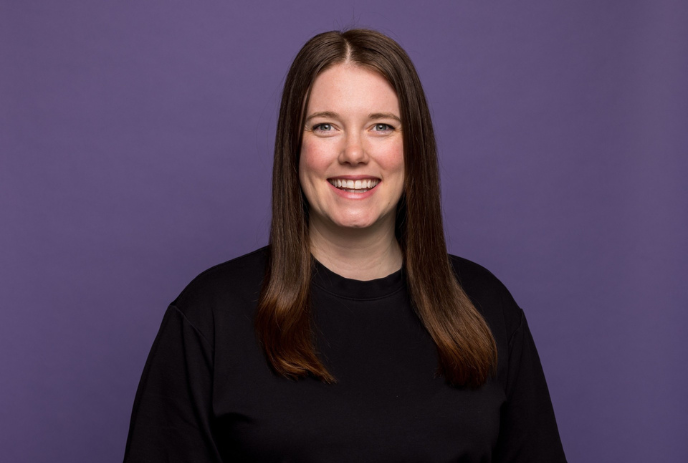 Smiling women artist with brown hair in black shirt, standing against solid purple background, facing camera.