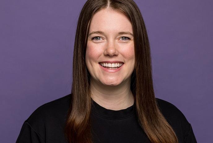 Smiling women artist with brown hair in black shirt, standing against solid purple background, facing camera.