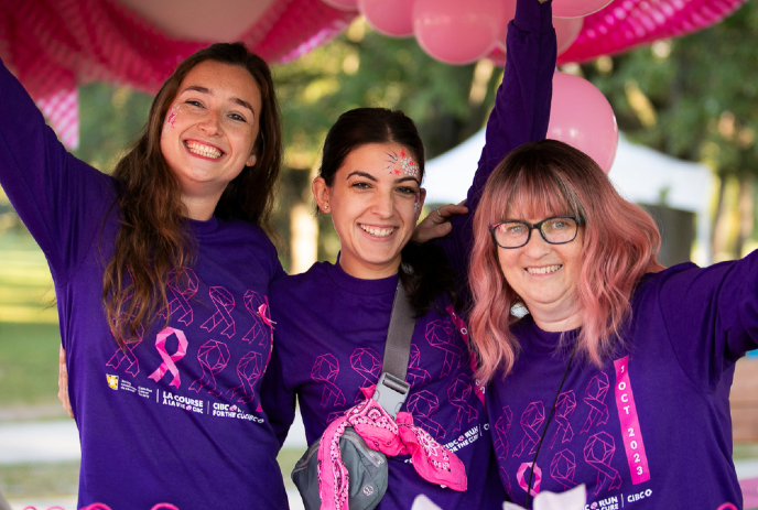Three smiling women in purple shirts with pink ribbons pose under balloons at a breast cancer awareness event.