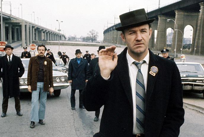 Man in dark coat gesturing on urban street with group of suited men and police near concrete overpass.
