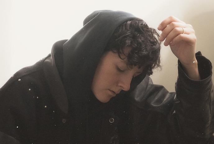 Artist in dark hoodie and jacket against a plain light backdrop, looking downwards.