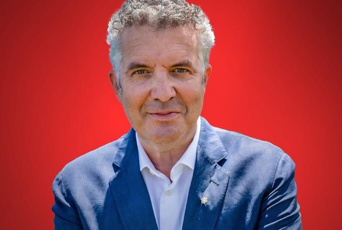 Rick Mercer in a blue suit stands arms folded in red background.