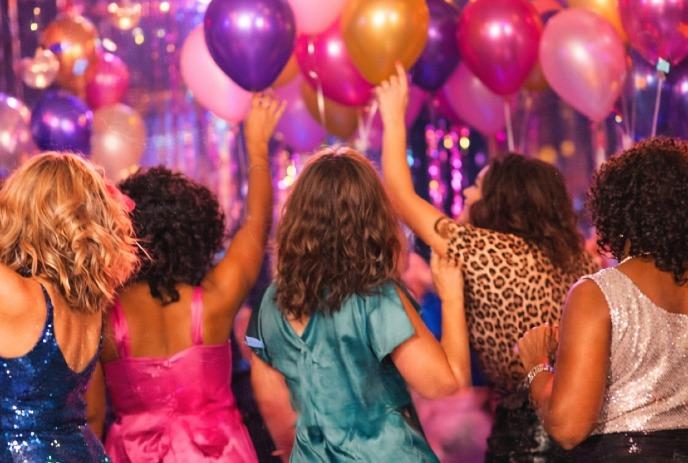 Women in vibrant dresses dance joyfully, surrounded by colorful balloons and shimmering lights.