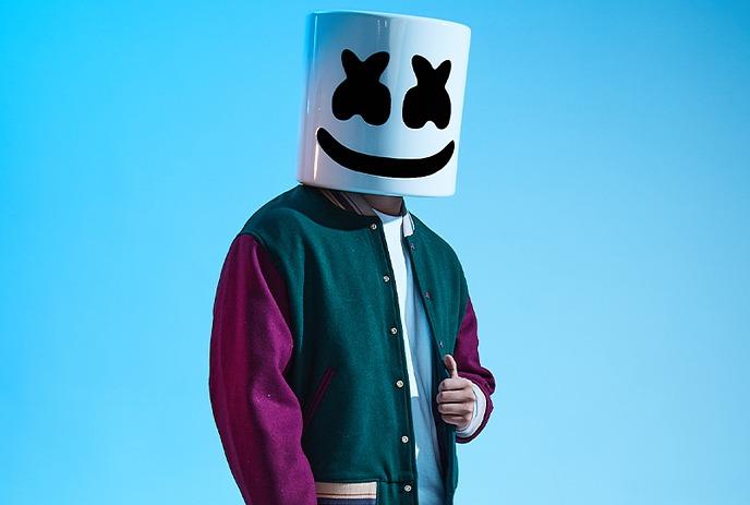 Musician Marshmello poses against a blue background, wearing his signature white helmet and a varsity jacket.