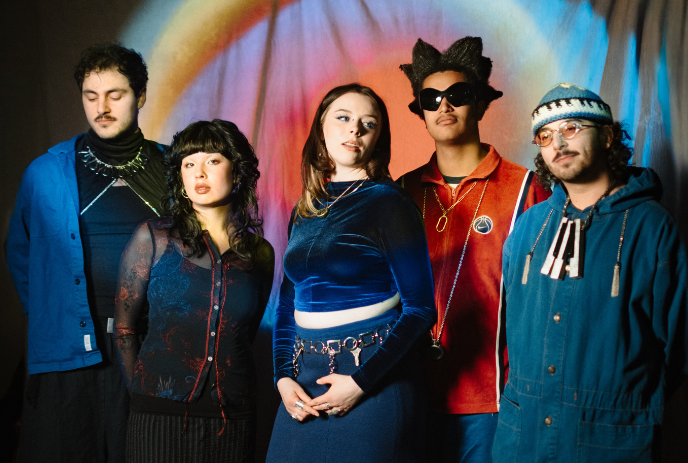 Five artists stand before a bright circular backdrop wearing bold colorful outfits in a vivid group pose.