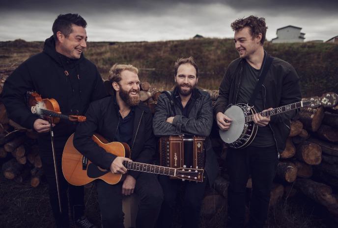 4 members of the band Rum Ragged, posing in a field with their instruments, smiling under a grey sky.