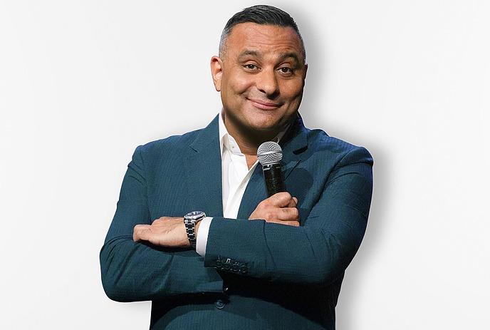 Russell Peters wearing a suit, posing for the camera against a white background.