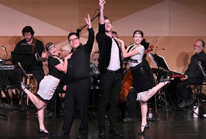 Performers pose on stage beside musicians creating lively theatrical scene.