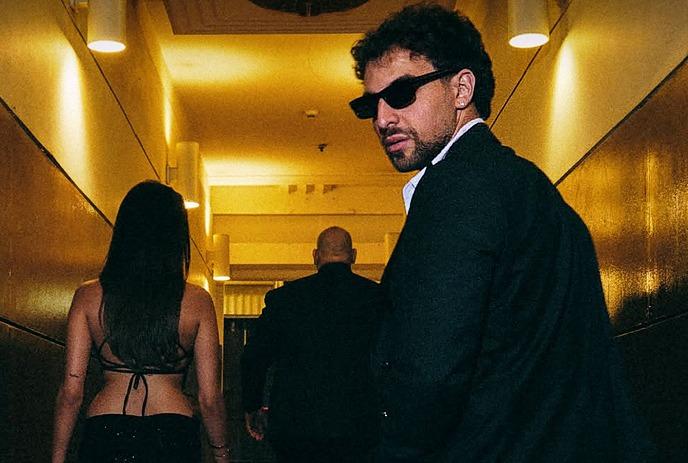 Saint Levant in dark suit and sunglasses walks a hotel hallway with two figures ahead on red carpet.