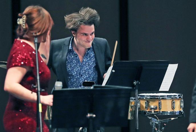 Drummer with flying hair plays snare beside singer in red dress, both on the music stag