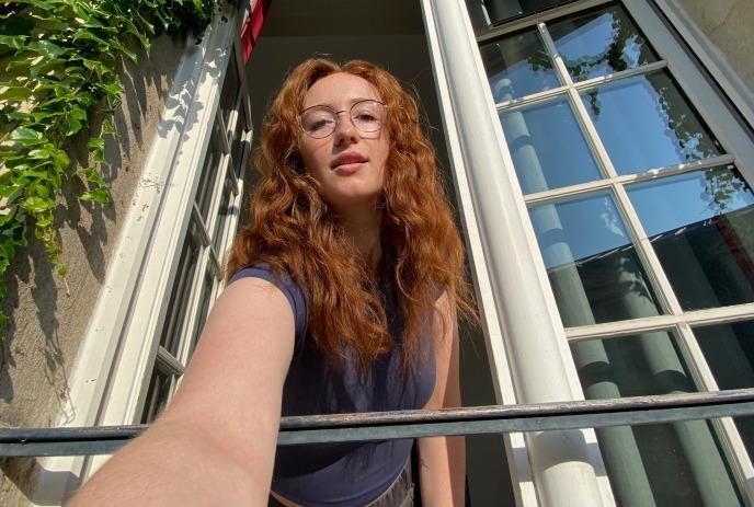 Artist Sam Peco leans from open window with long hair, glasses and ivy on wall in bright daytime light.