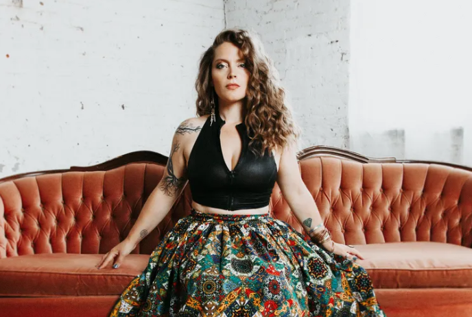 Sandra Bouza seated on a vintage rust-colored sofa, wearing a black top and patterned skirt indoors.