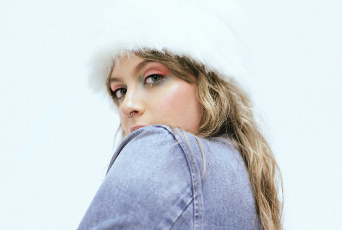 Arists in fluffy white hat and denim jacket looks back with bright eye makeup against plain backdrop.