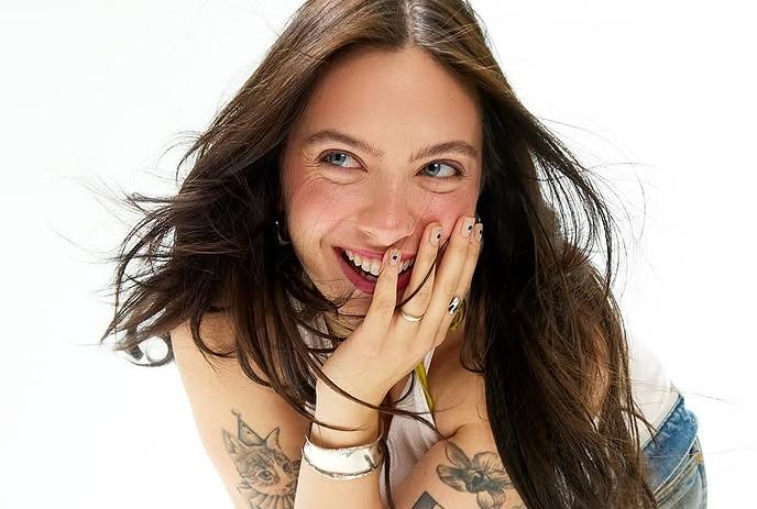 Saveria laughing with hands near her face, showing tattooed arms and casual attire.