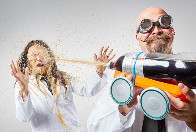 Two people in lab coats react as soda sprays wildly from a bottle on a small, wheeled stand.