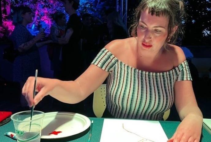 Angie Quick paints at a table with a crowd in the background under colorful lights.