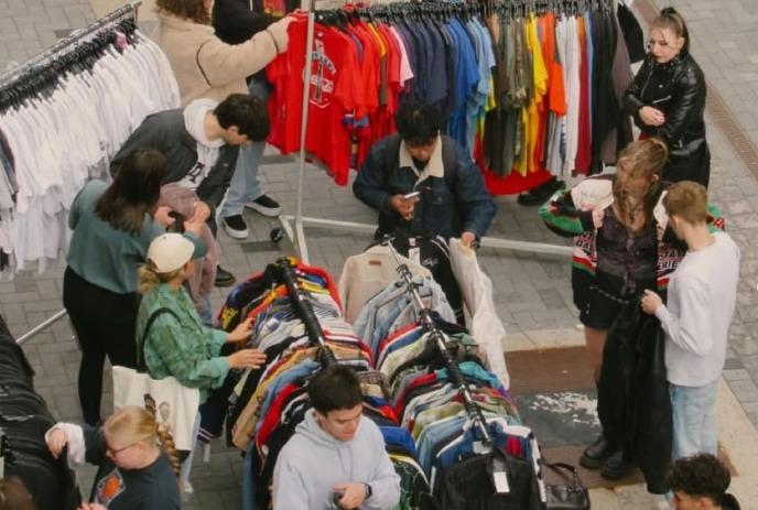 Outdoor thrift market with people browsing colorful clothes on racks in a casual, communal street setting.