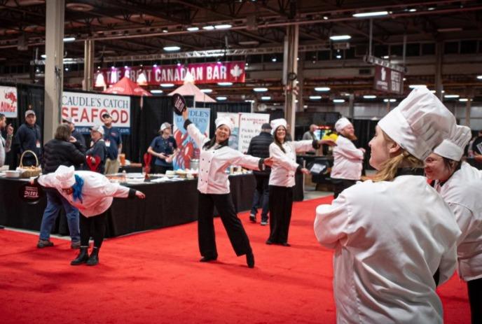 A group of chefs in white uniforms joyfully dancing on a red carpet at a bustling food event.