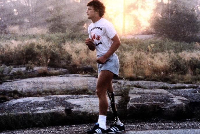 Terry Fox runs on a rocky path with a prosthetic leg, wearing a marathon T-shirt and shorts at sunset.