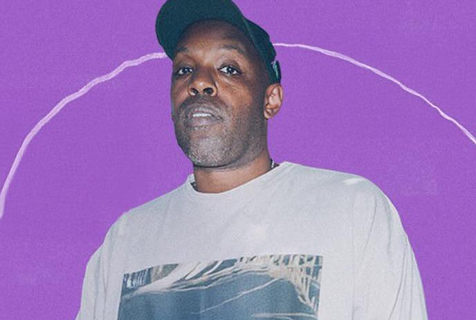 Shad wearing a black shirt and black hat, wearing a silver chain and standing against a green wall.