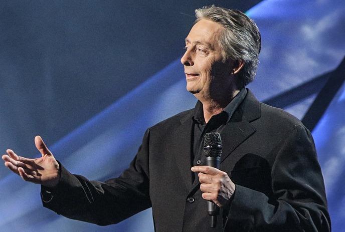 A speaker in a black suit holds a microphone and gestures on a blue-lit stage with dramatic light beams in the background.