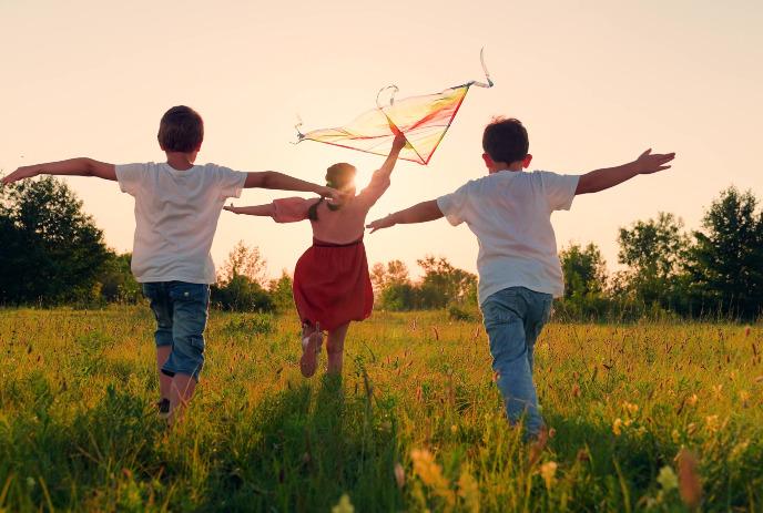 Children run through warm sunset field as bright kite lifts above grass in joyful playful golden motion.