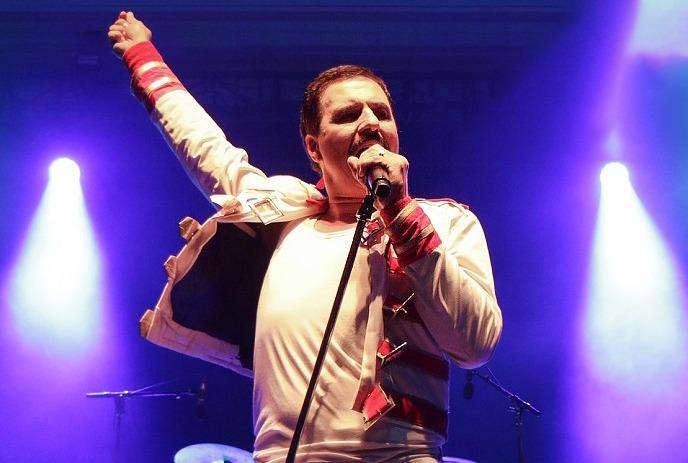 Person dressed as Freddie Mercury with yellow shirt and jacket singing into a microphone.