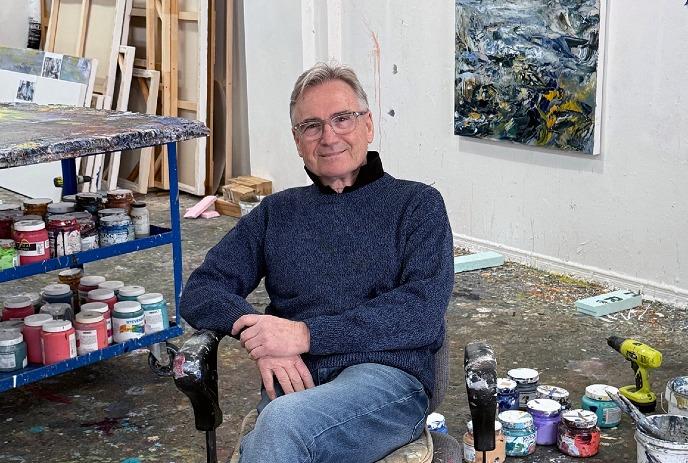 Michael Smith in a cozy sweater sits contentedly in an art studio, surrounded by colorful paint jars and artworks.
