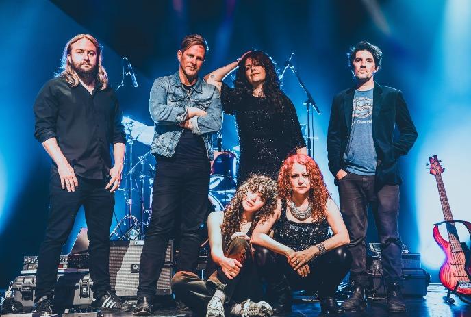 A group of six band members poses confidently on a dimly lit stage with blue lighting and instruments.