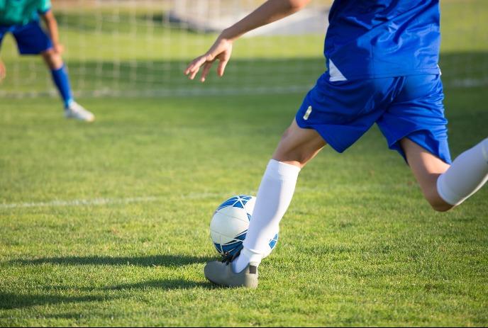Player in blue kicks a soccer ball on grass as another player waits near the goal in the background.