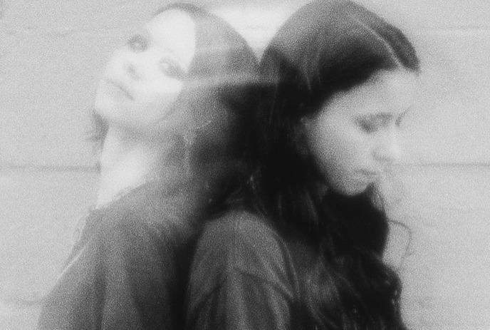 Two female members of the band Softcult stand back-to-back against a brick wall in a grainy black-and-white photo.