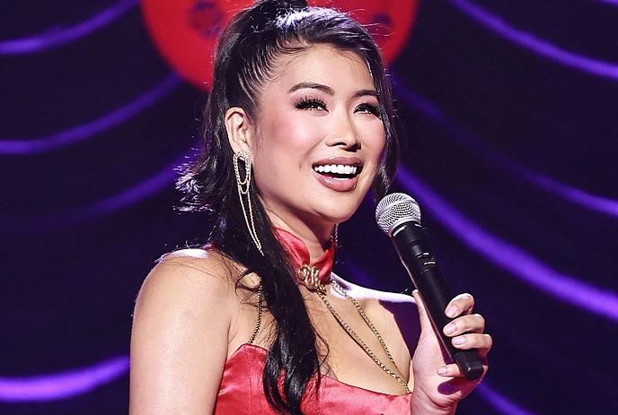 Smiling performer in a red satin dress holding a microphone on stage, with elegant earrings and purple lighting