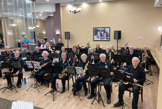 An adult swing band in black attire sits with saxophones and trombones, smiling in a bright hall, ready to play.